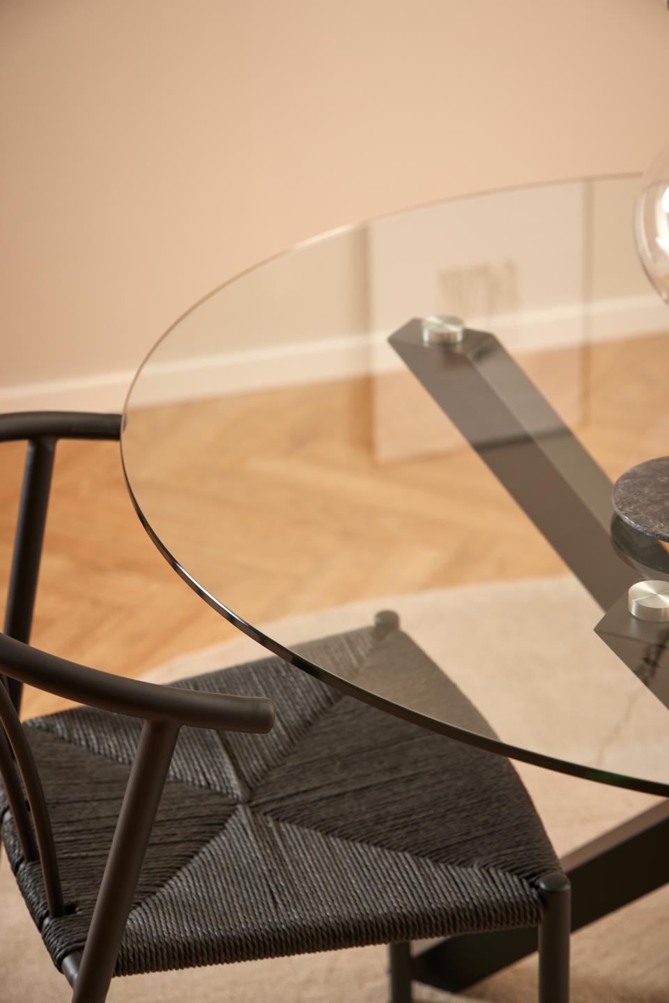 Close-up of a modern dining table with a clear oval glass top and dark metal frame, paired with a woven dark gray chair in a warm beige room.