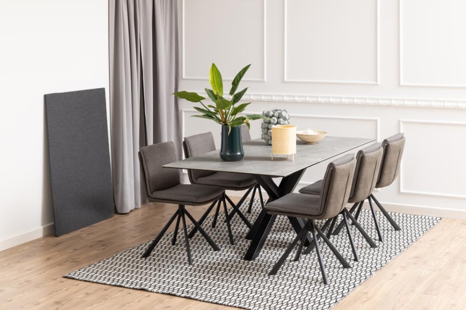 A modern dining set featuring a rectangular gray stone-look table with a black metal X-frame base, surrounded by six gray upholstered swivel chairs. The set is styled on a black and white patterned rug in a bright dining room with light wood floors, gray curtains, and white wall paneling.