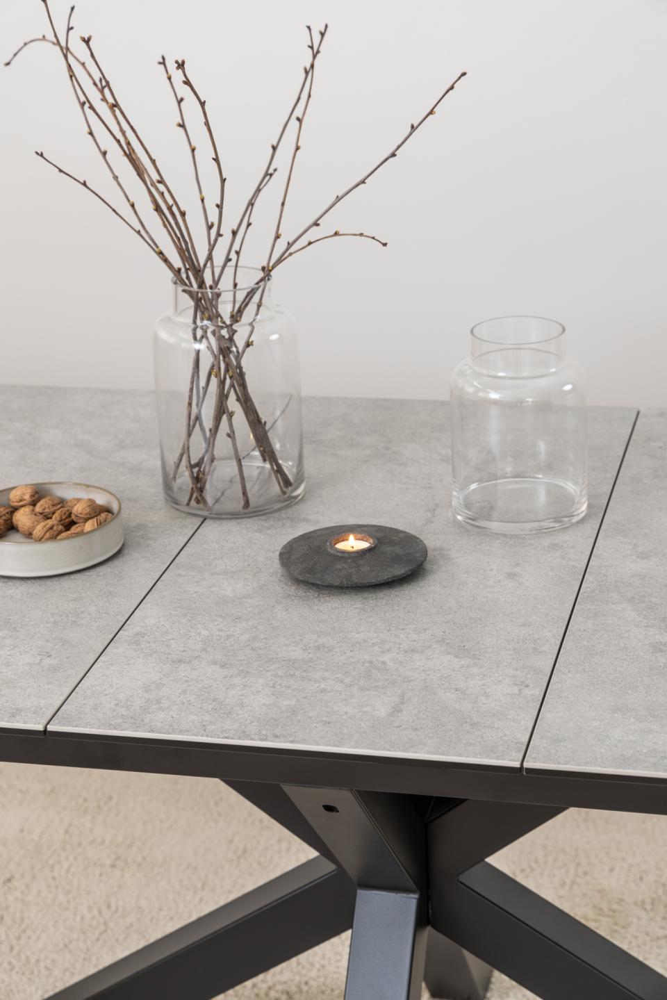 A dark round stone candle holder with a lit tealight candle sits on a gray extending dining table, styled with a glass vase of branches and a clear glass jar.