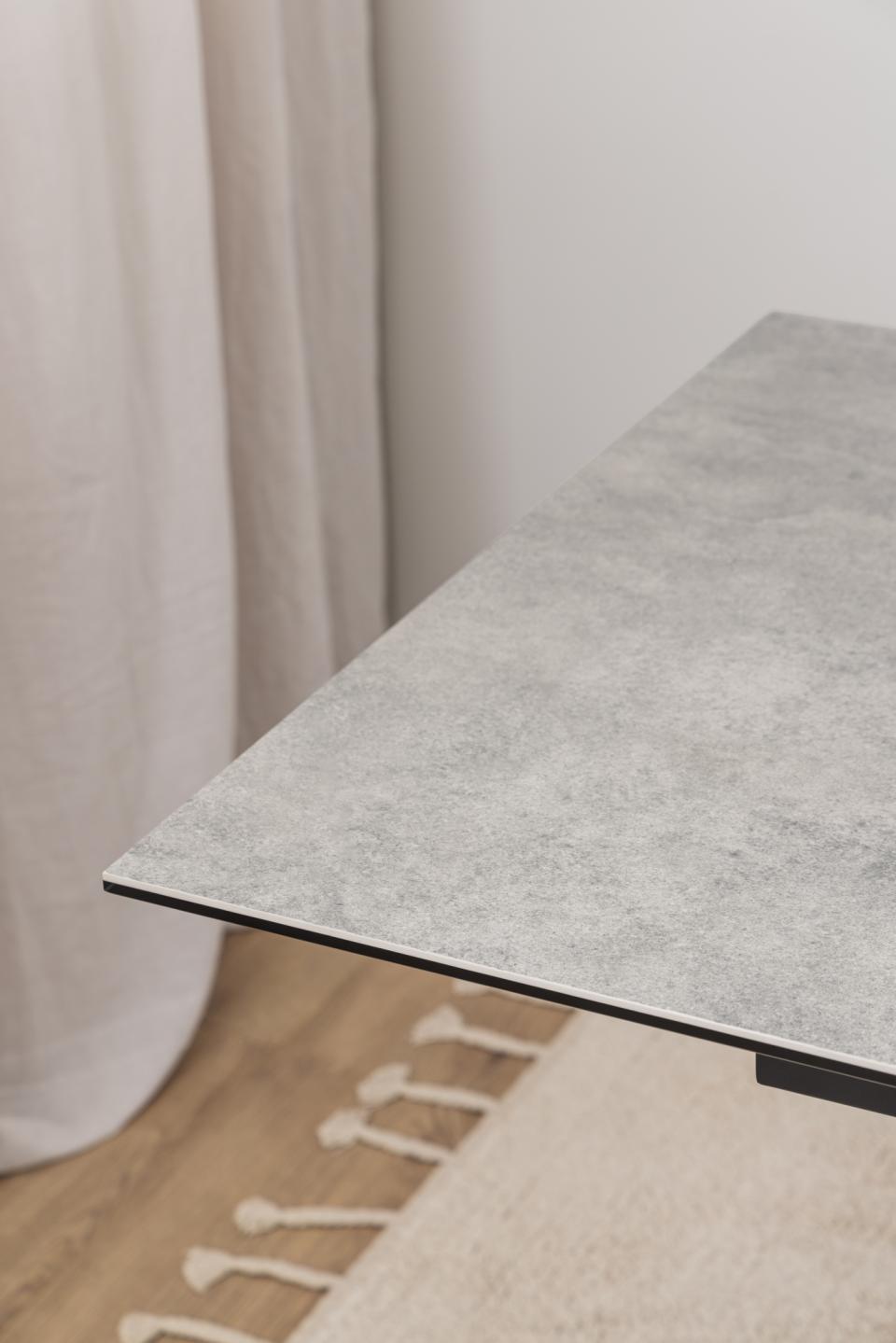 A close-up of a gray ceramic tabletop extending over a light wood floor, with a cream rug and neutral wall and curtain in the background.