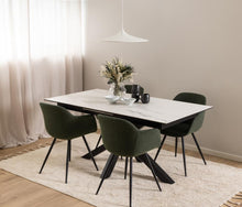Load image into Gallery viewer, A modern dining table set in a bright, minimalist room. The set includes a rectangular white marble-look table with a black metal frame and four dark green upholstered dining chairs with black legs.
