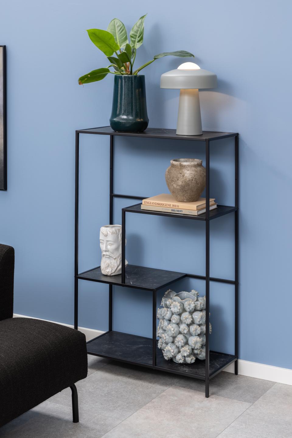 A black metal open shelving unit styled with home decor, including a potted plant, a table lamp, books, and decorative vases, set against a blue wall.