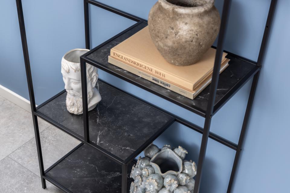 A black metal shelving unit with black marble-look shelves, styled with a textured stone pot, books, a white sculptural vase, and a gray ceramic vessel against a blue wall.