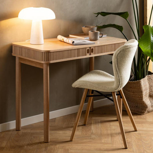A rectangular oak veneer desk with concealed drawers and tambour doors, featuring a decorative slatted element, placed in a room with a lamp and a chair.