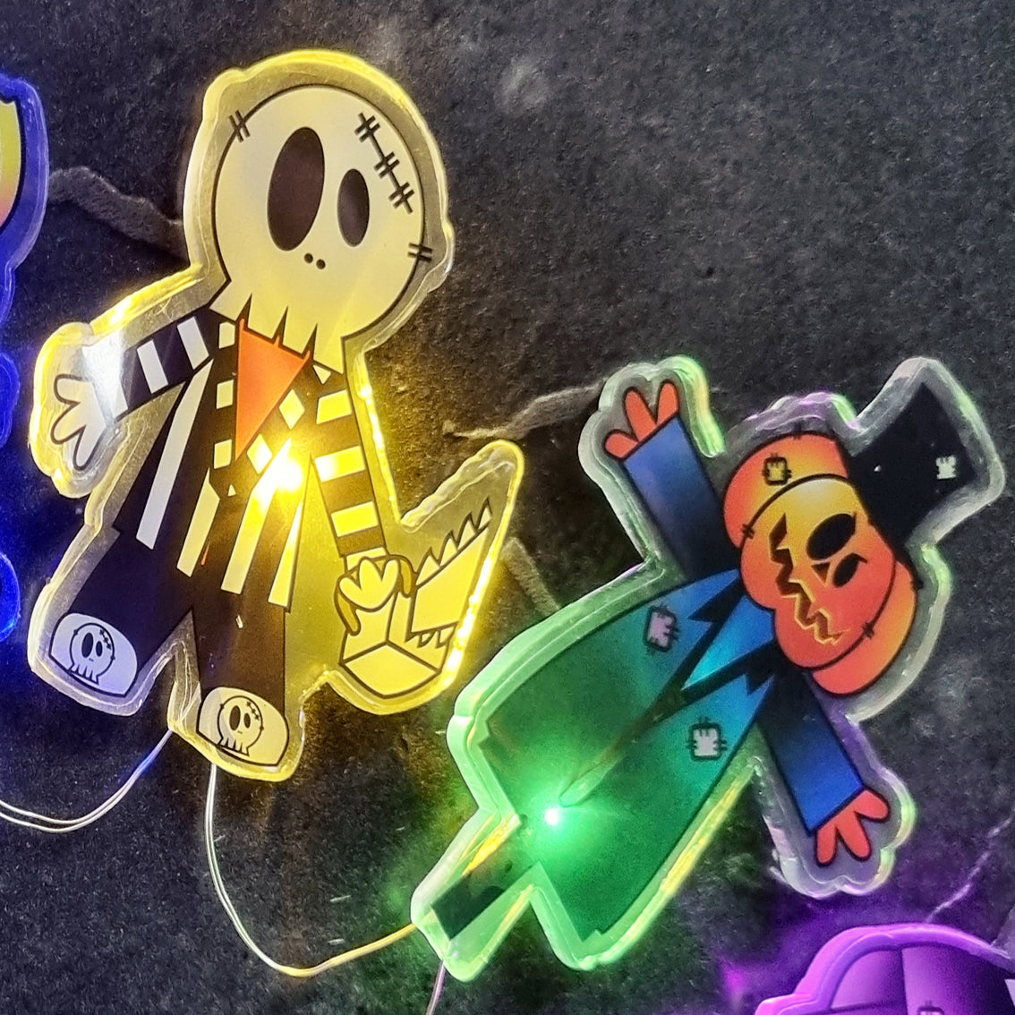 Two Halloween-themed skeleton figurines with glowing LED lights, one in a yellow-and-black striped suit and one in a blue-and-orange suit, displayed on a dark textured surface.