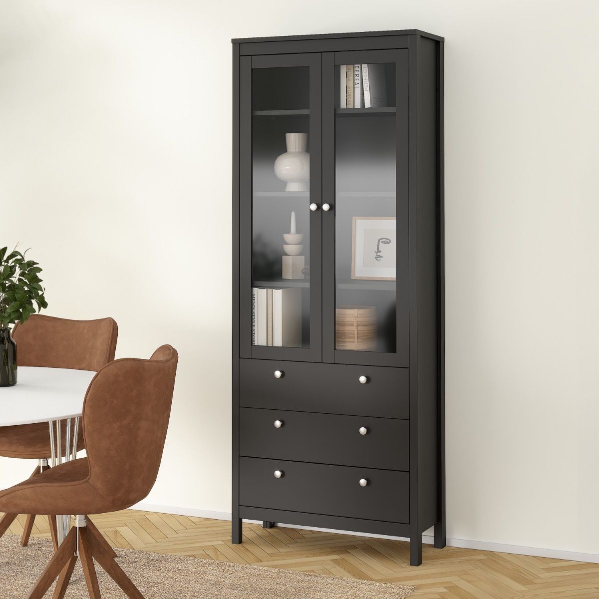 A tall black wooden display cabinet with two glass upper doors and four drawers below, shown in a styled dining room setting next to a table and chairs.