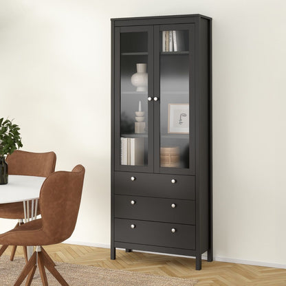 A tall black wooden display cabinet with two glass upper doors and four drawers below, shown in a styled dining room setting next to a table and chairs.