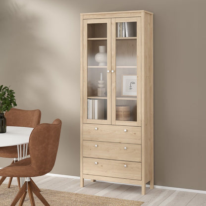 A brown light oak Madrid cabinet with two glass doors, three drawers and three shelves filled with home accessories and standing against a beige wall in a modern home with a table and chairs