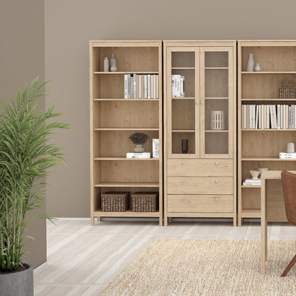 A brown light oak Madrid cabinet with two glass doors, three drawers and three shelves in the middle of matching shelving units in a modern room with a rug, plant and a table and chair just viewable