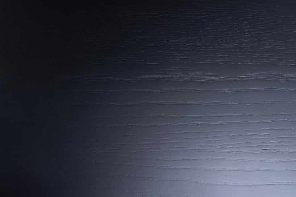 A close-up of a dark wood surface with visible grain texture and a lighter highlight near the bottom right.