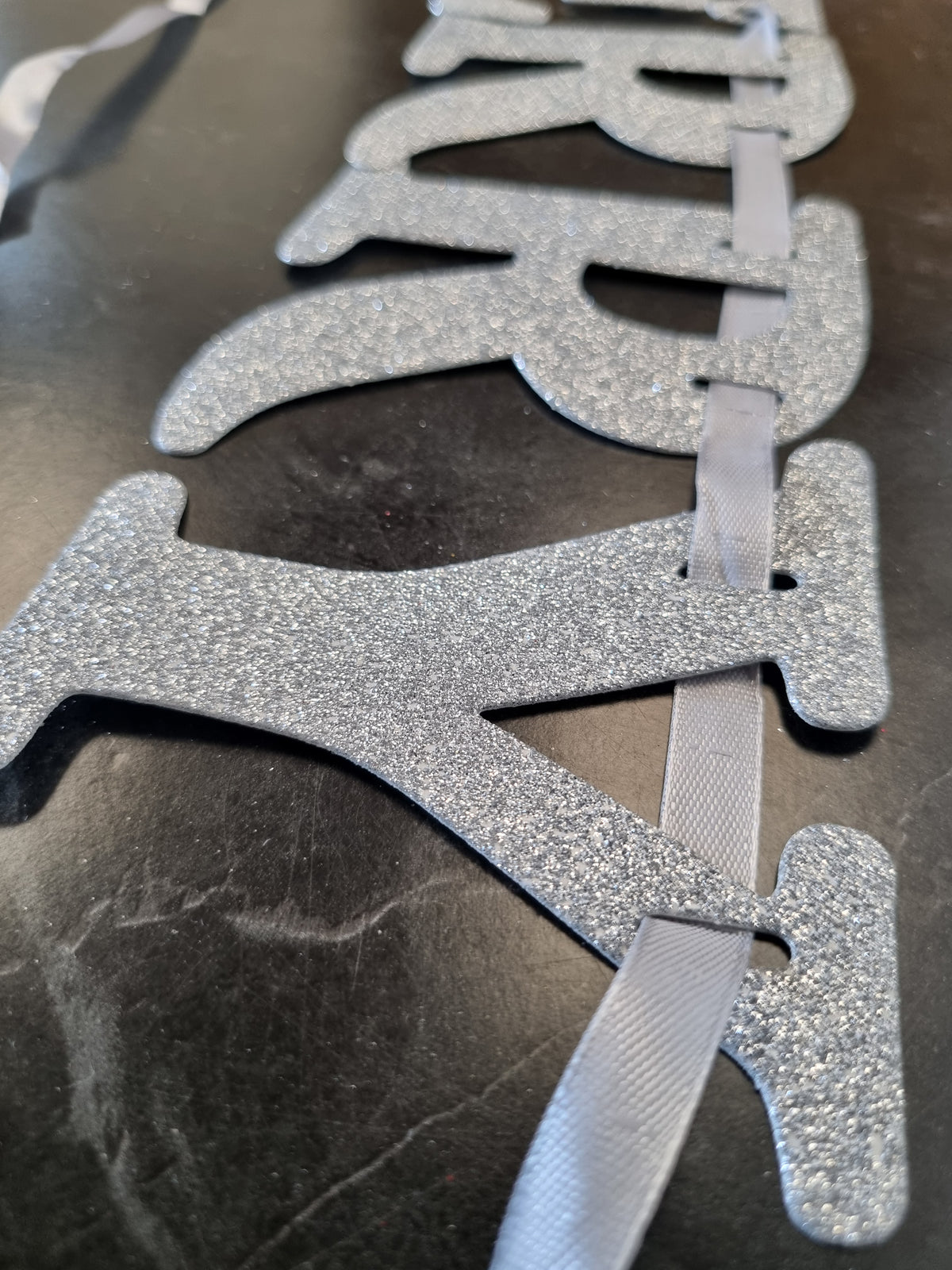 Silver glitter letter banner spelling 'HAPPY' with gray ribbon loops, shown close-up on a dark surface.