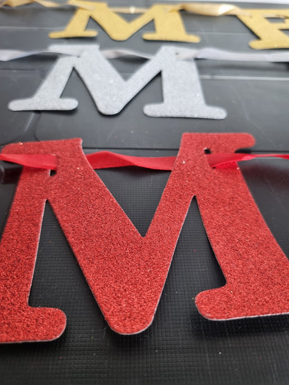 A close-up of a glittery red letter 'M' with a red ribbon, with other glittery letters in gold and silver partially visible in the background on a dark textured surface.