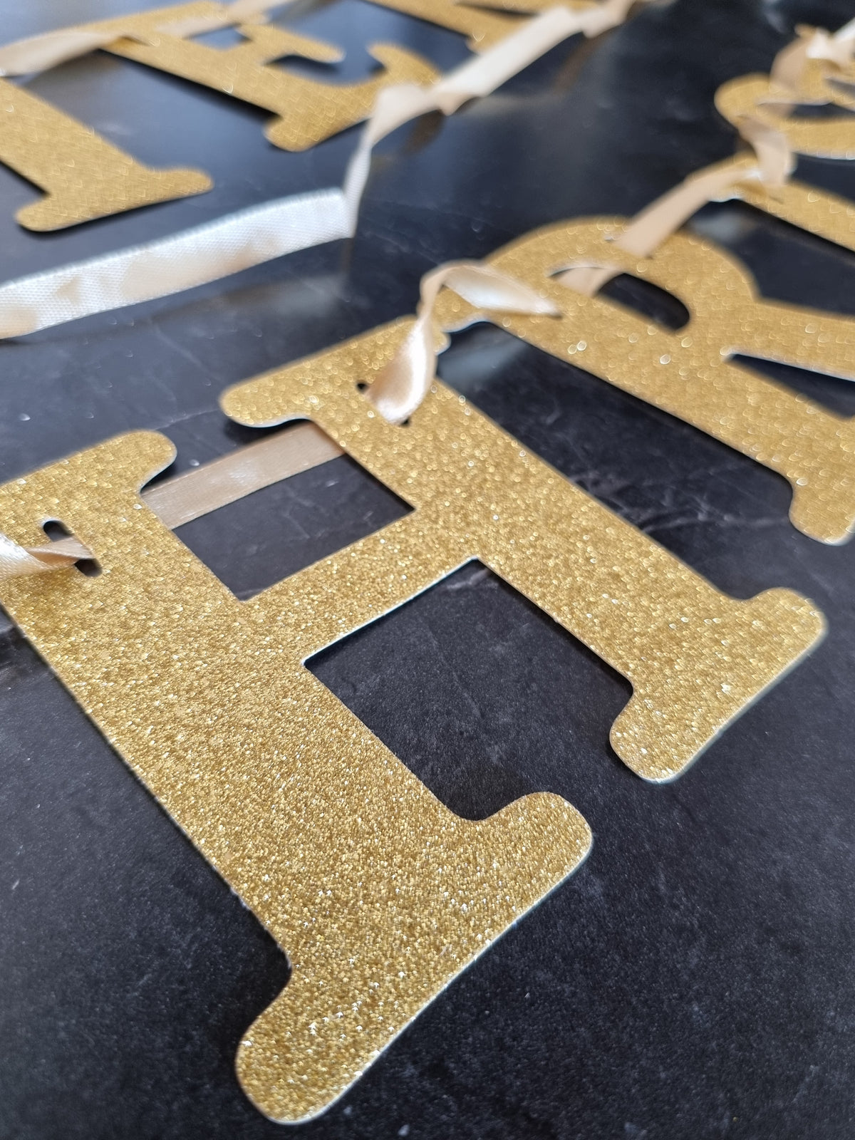 Gold glitter letter banner garland with hanging ribbon, shown close up on a dark surface.