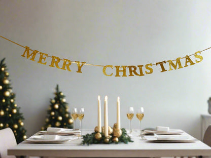 A gold glitter 'MERRY CHRISTMAS' banner hangs across a dining room wall above a set dining table with white plates, folded napkins, lit taper candles, and wine glasses. Two decorated Christmas trees are visible in the background against a light gray wall.