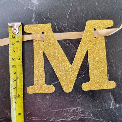 A glittery gold letter 'M' monogram decoration with a beige ribbon, shown next to a yellow tape measure on a dark marble surface.