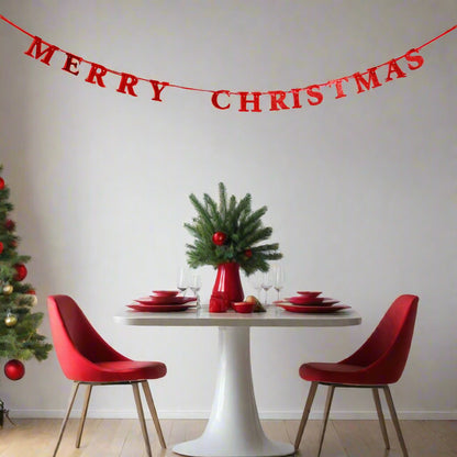 A festive holiday dining room scene with a red 'MERRY CHRISTMAS' banner hanging across a white wall above a white table set with red chairs, red plates, and a green pine vase centerpiece.