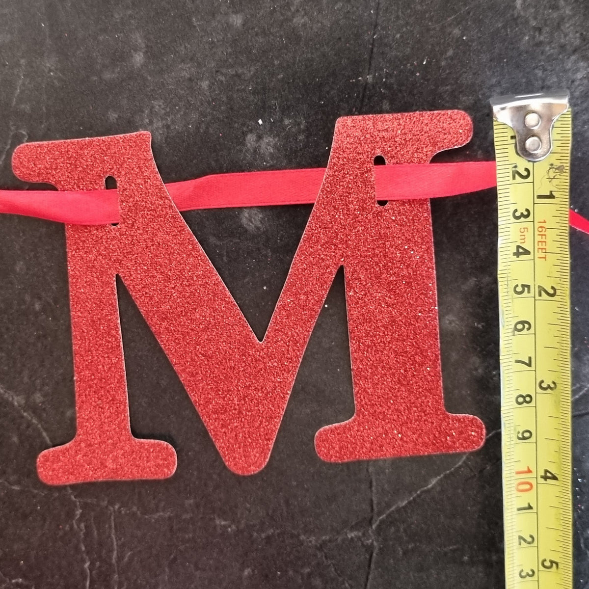 A red glittery letter 'M' hanging from a red ribbon, shown next to a yellow tape measure on a dark textured surface.