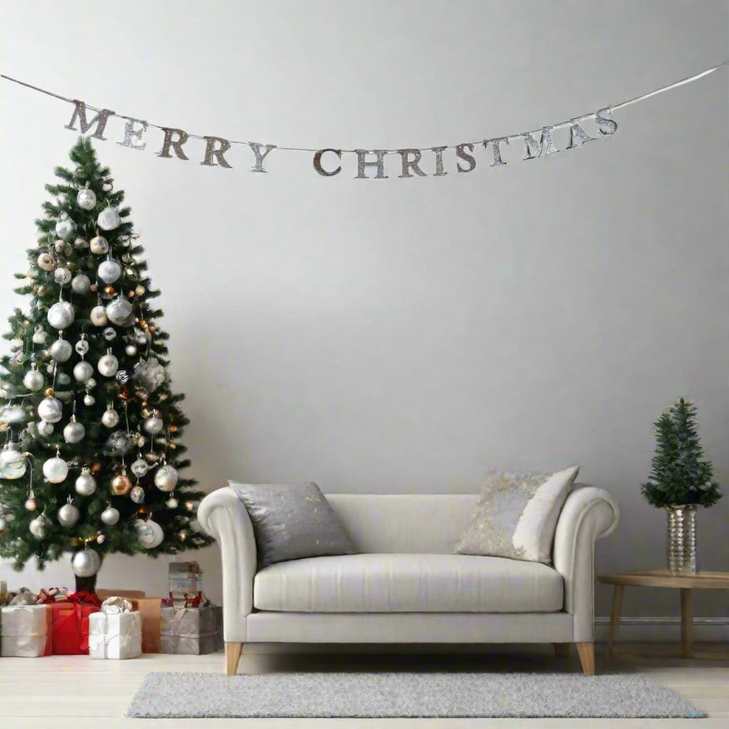 A festive holiday living room scene featuring a silver 'MERRY CHRISTMAS' banner garland strung across a light gray wall above a cream-colored sofa, next to a decorated Christmas tree and a small potted tree on a side table.