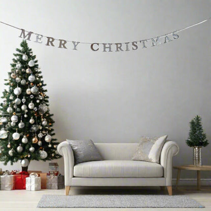 A festive holiday living room scene featuring a silver 'MERRY CHRISTMAS' banner garland strung across a light gray wall above a cream-colored sofa, next to a decorated Christmas tree and a small potted tree on a side table.