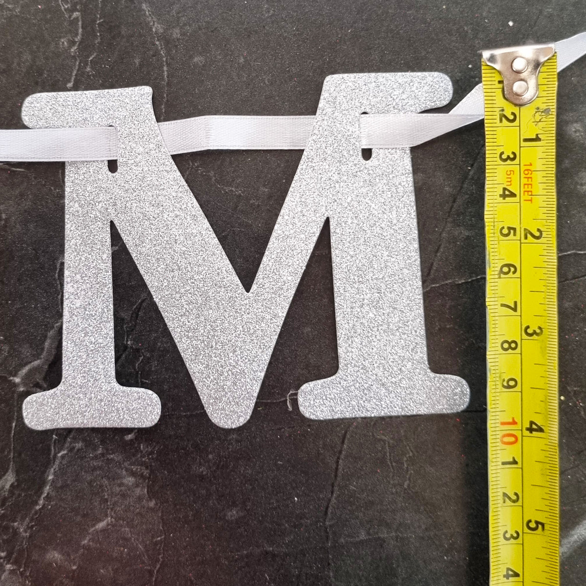 A glittery silver letter 'M' hanging from a white ribbon, shown next to a yellow measuring tape on a dark marble surface.