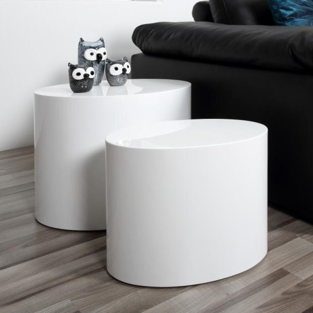 Two white high gloss tables of different sizes, with the smaller table fitting inside the larger one, placed on a wooden floor against a black sofa background with decorative mice figures on top.