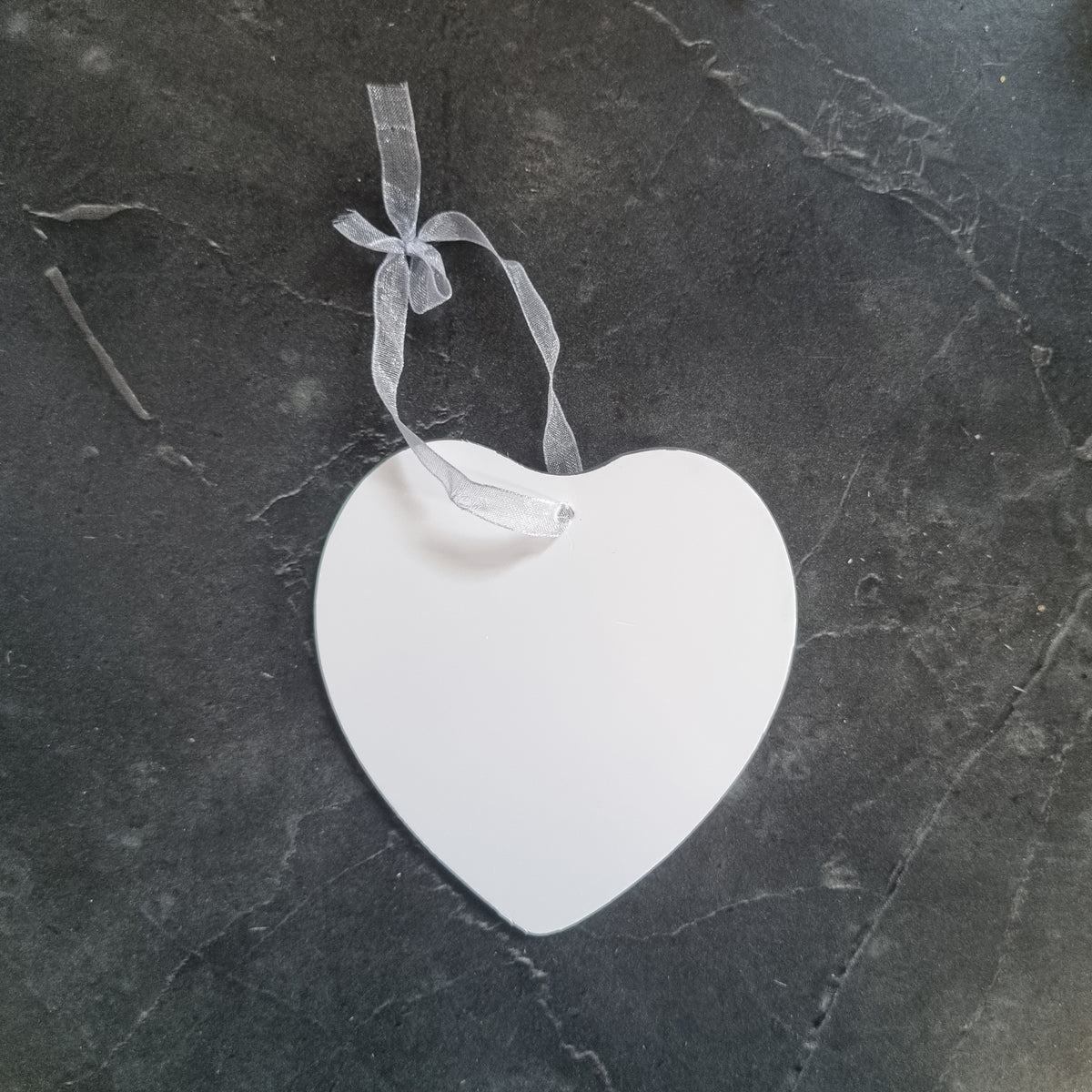 A blank white heart-shaped ornament with a sheer white ribbon loop, placed on a dark gray textured stone surface.