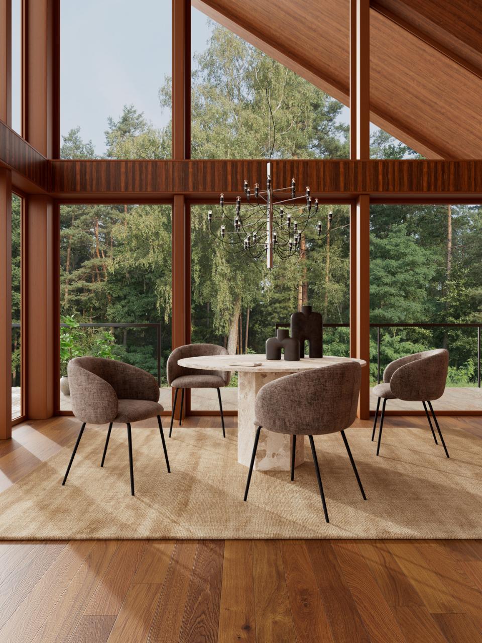 A modern dining room scene with a round white table and four upholstered gray chairs arranged on a beige rug, set inside a wood-framed room with large windows overlooking a forest.