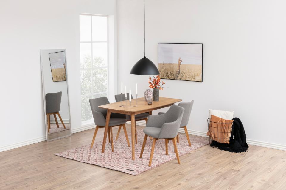 A modern dining room setup featuring a light wood rectangular table with six gray upholstered chairs, a black pendant light, a pink patterned rug, and a tall floor mirror reflecting the scene.