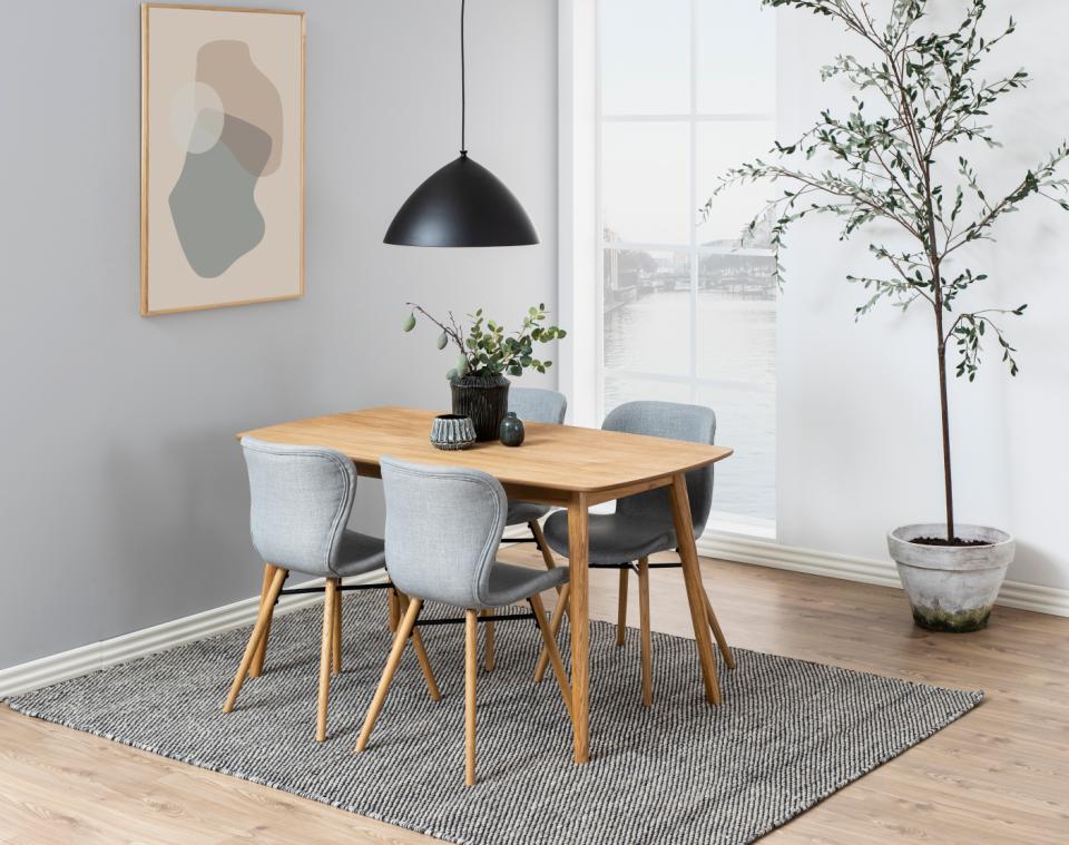 A modern dining set featuring a light oak wood rectangular table and four gray upholstered chairs with wooden legs, styled in a bright, contemporary dining room with a black pendant light, gray rug, and potted tree.