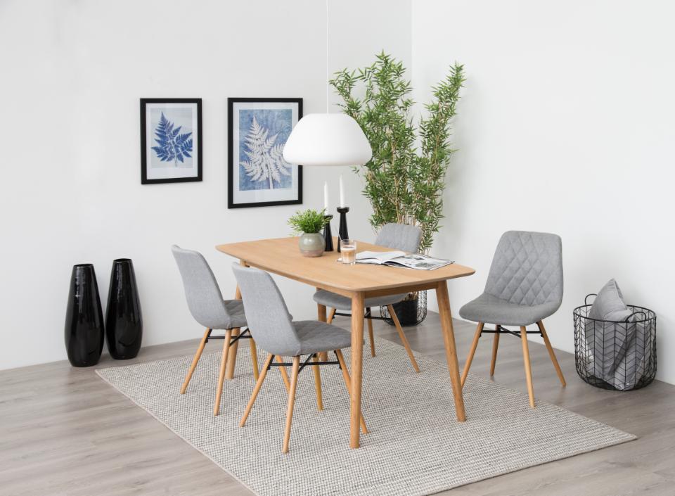 A modern dining room setup featuring a rectangular light oak wood table surrounded by four gray upholstered dining chairs with wooden legs, set on a textured light gray area rug. The room has white walls, two black decorative vases, framed botanical prints, a white pendant light, and a potted bamboo plant.