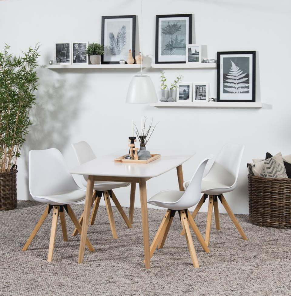 A modern dining set featuring a white rectangular table with light wood legs and four matching white molded chairs with angled wooden legs, styled in a bright, minimalist dining room with white shelves, framed art, and a potted plant.