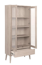 Load image into Gallery viewer, A tall light oak wood armoire cabinet with two glass doors, one of which is open to reveal four interior shelves, and a drawer pulled out at the bottom, shown on a white background.
