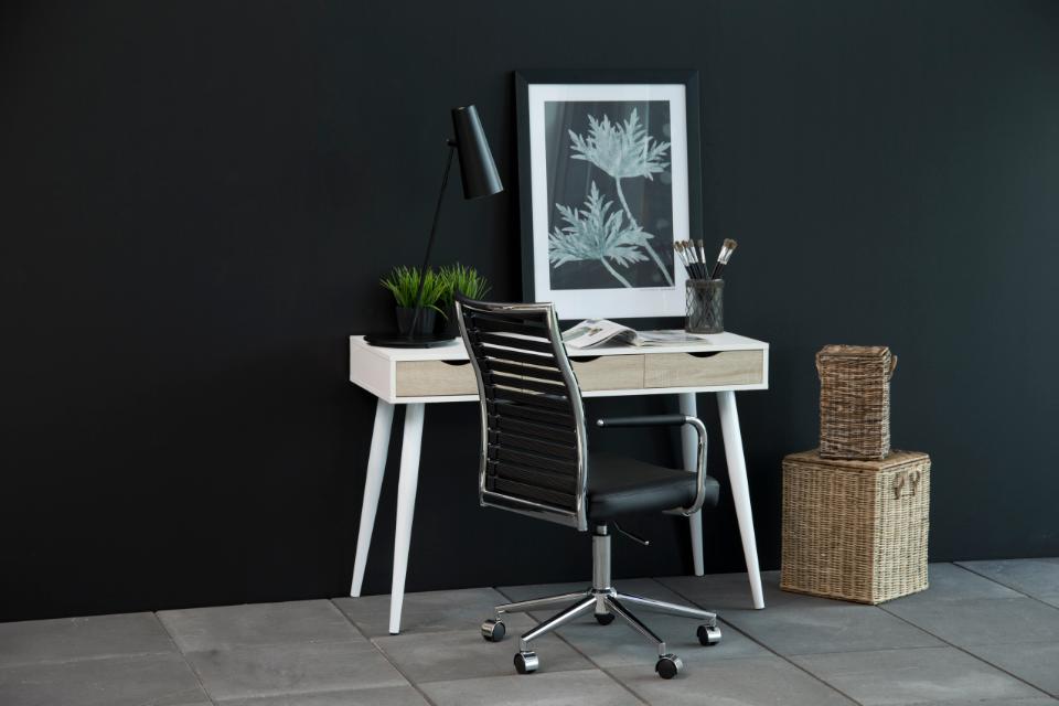 A modern home office setup featuring a light wood and white desk with tapered legs, a black slatted office chair, a black desk lamp, and a framed botanical print against a dark charcoal wall.