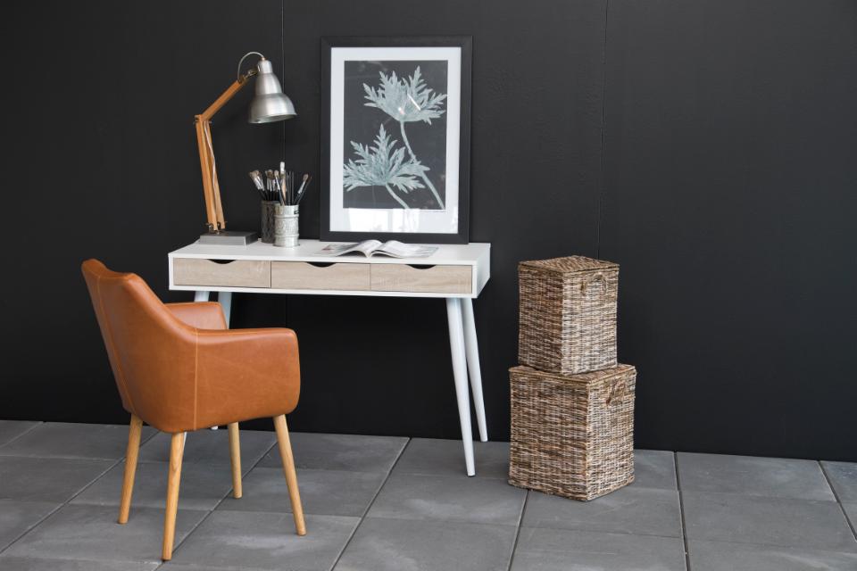 A styled interior scene with a white writing desk against a dark charcoal wall, a tan leather chair, a silver desk lamp, a framed botanical print, and two stacked woven storage baskets on a gray tiled floor.