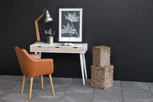 Load image into Gallery viewer, A styled interior scene with a white writing desk against a dark charcoal wall, a tan leather chair, a silver desk lamp, a framed botanical print, and two stacked woven storage baskets on a gray tiled floor.
