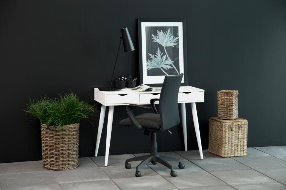 A modern home office setup featuring a white desk with tapered legs, a black swivel office chair, a black desk lamp, and a framed botanical print on a dark charcoal wall. Decorative wicker baskets and a potted green plant complete the scene on a gray tiled floor.