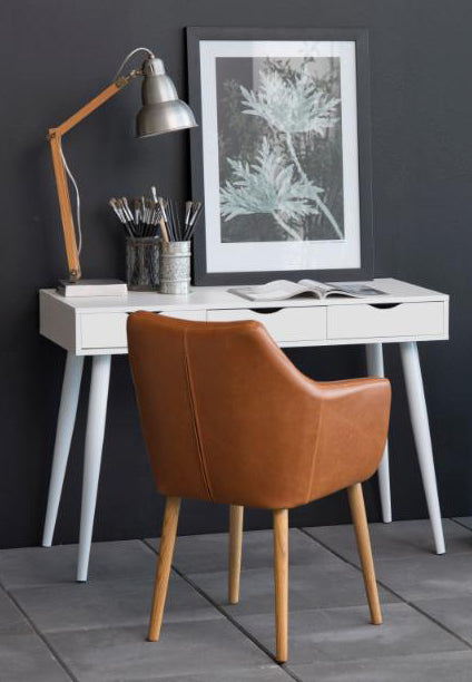 A modern home office setup featuring a white writing desk with tapered legs, a brown leather armchair, a silver adjustable desk lamp, and a framed botanical print against a dark gray wall.