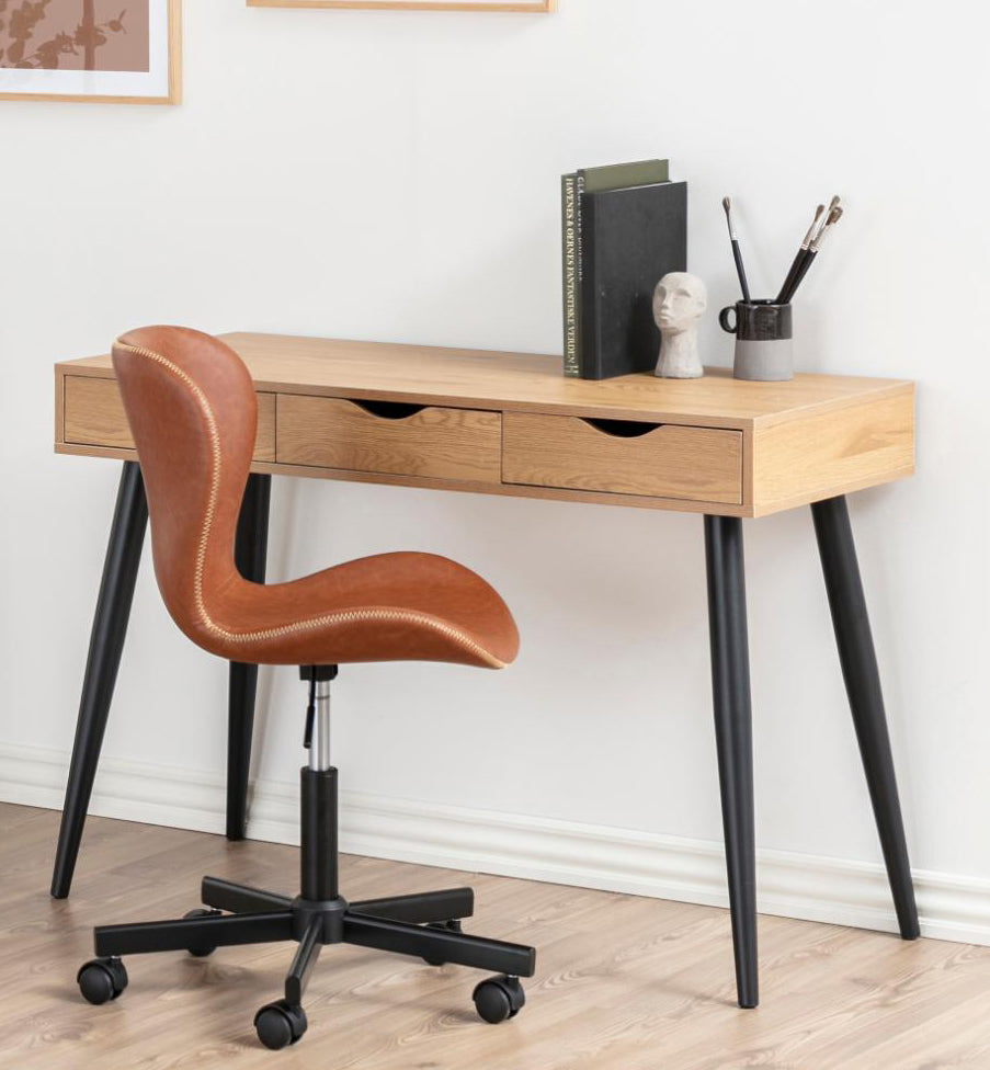 A mid-century modern writing desk with a light oak wood top, three drawers, and black tapered legs, paired with a brown leather swivel chair in a bright home office setting.