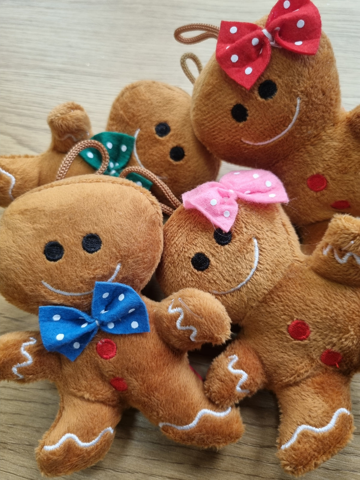 A group of small plush gingerbread person toys arranged together on a light wood surface. The toys are brown with embroidered white icing details, black eyes, and smiling faces, and each wears a different fabric bow in red, green, pink, or blue with white polka dots.
