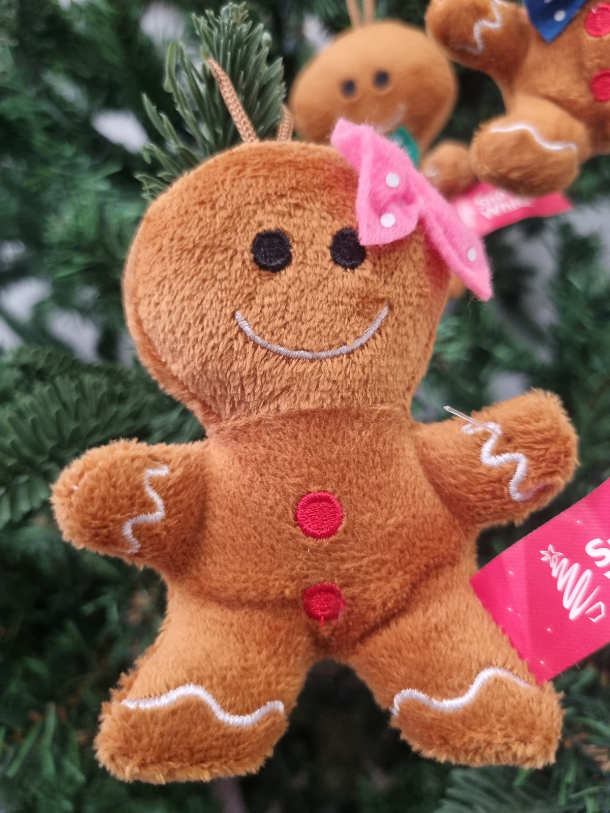 A plush gingerbread man Christmas ornament hanging on a green Christmas tree. The ornament is brown with white icing details, a pink bow, and red buttons, with a pink hang tag attached.