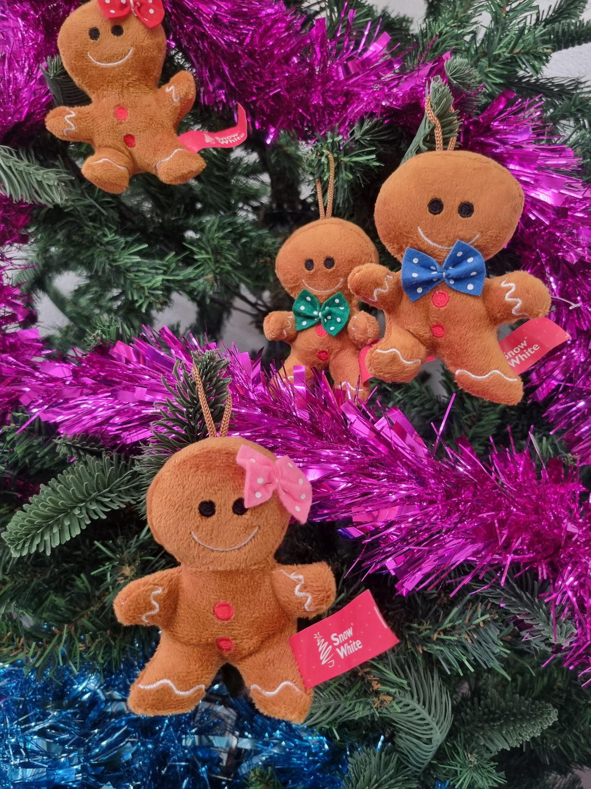 Four plush gingerbread man Christmas ornaments with colorful bows and red buttons, hanging on a decorated Christmas tree with bright pink and blue tinsel.