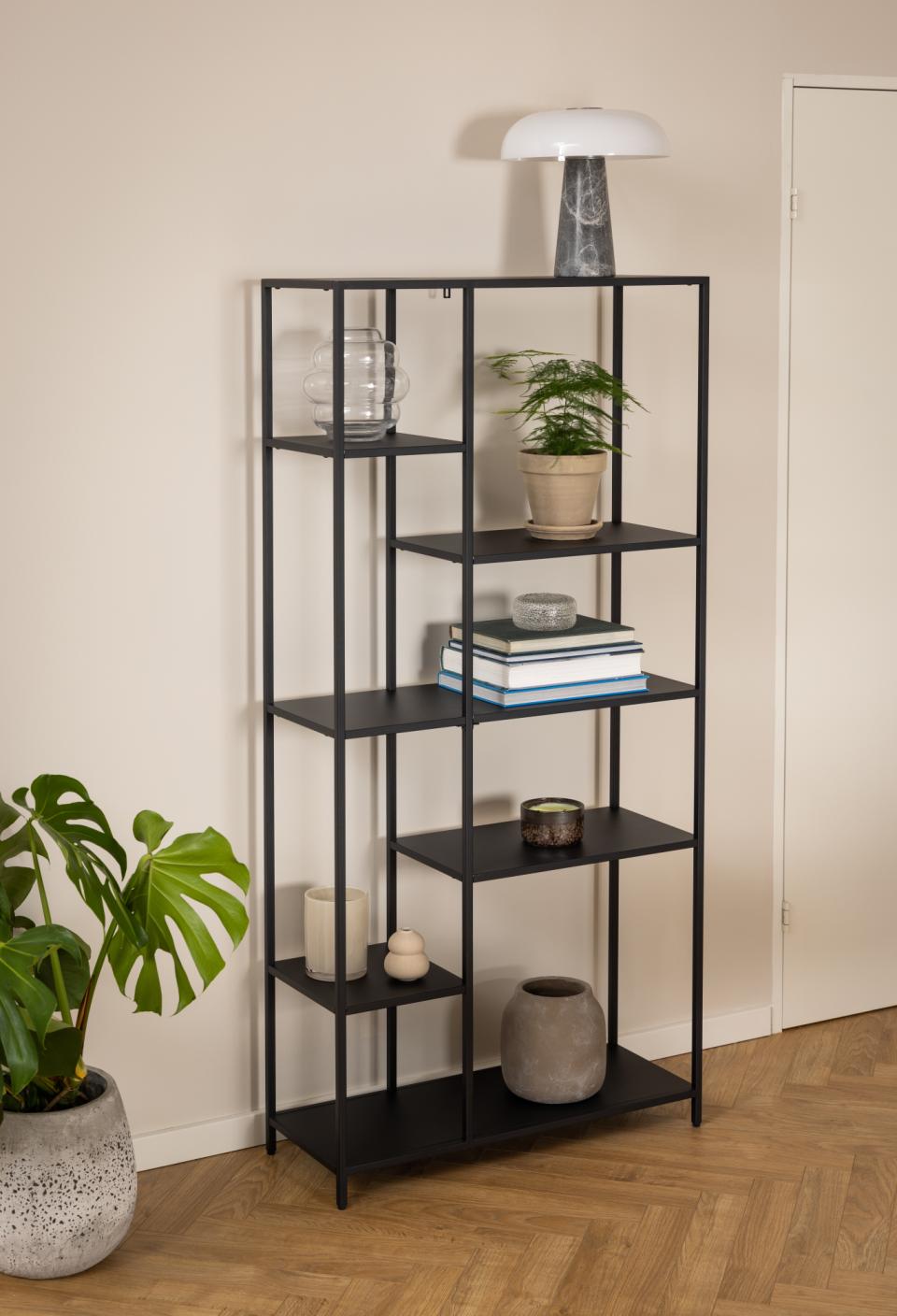 A tall black metal shelving unit with open shelves stands in a room corner against a beige wall and wood floor, styled with books, vases, a small plant, candles, and a lamp.