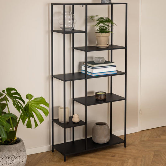 A black metal bookcase with six shelves, arranged in an asymmetrical design, displaying various decorative items and books. The bookcase is positioned in a room with a wooden floor and a plant to the side.