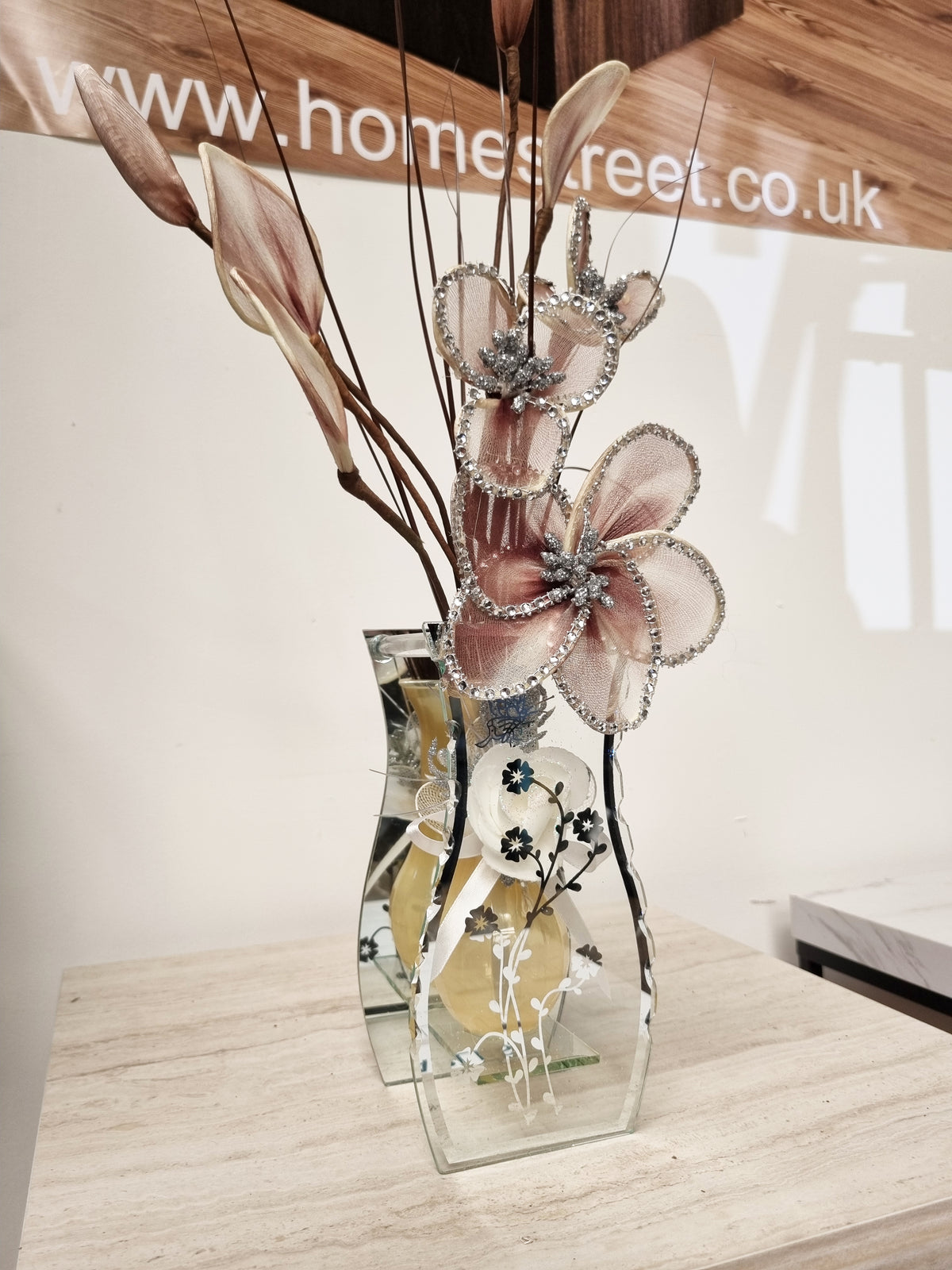 A decorative floral arrangement in a clear glass vase with mirrored edges, featuring large dusty pink flowers with silver glitter trim and dried brown stems, displayed on a light wood surface.