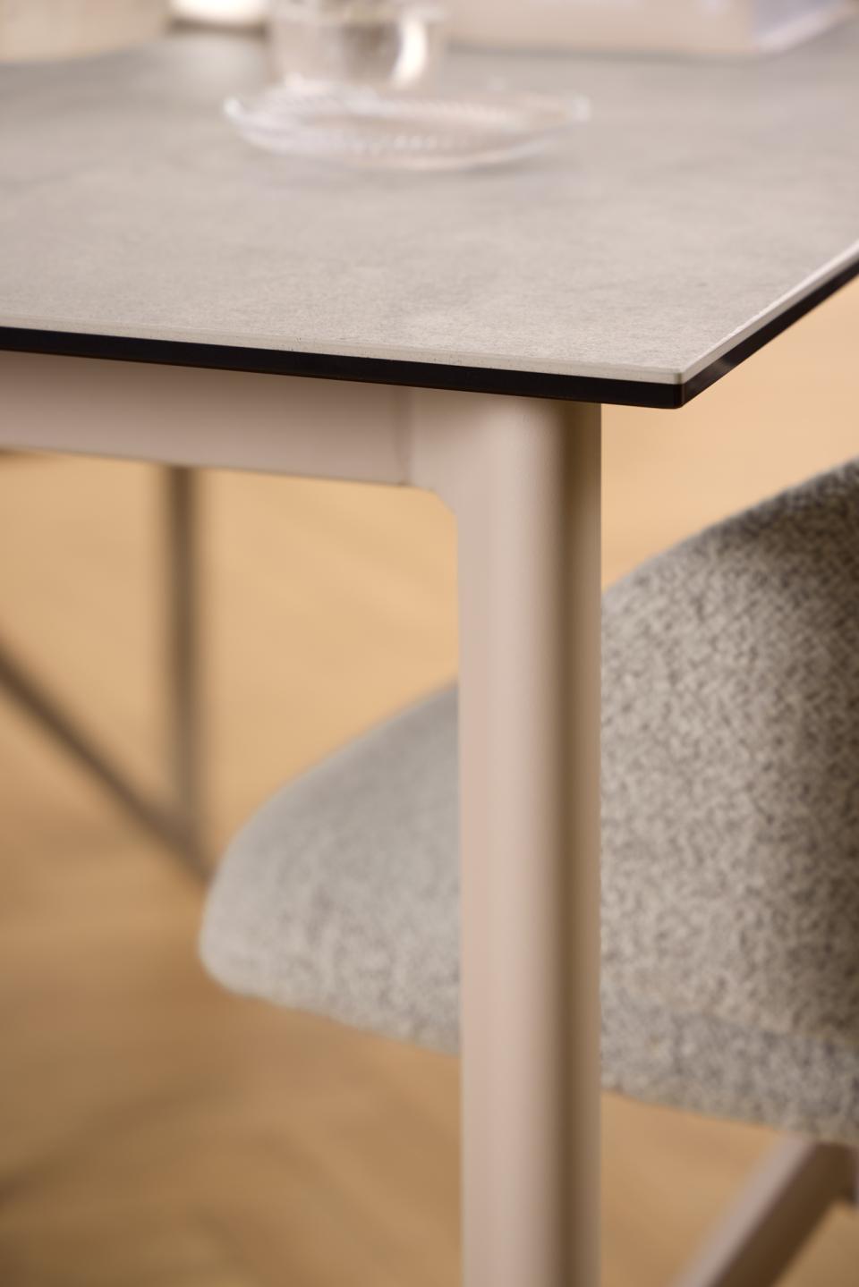 Close-up of a modern dining table with a light gray stone-look top and pale grey legs, shown alongside a textured light gray upholstered chair in a softly lit interior.