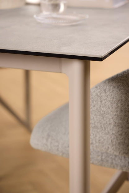 Close-up of a modern dining table with a light gray stone-look top and pale grey legs, shown alongside a textured light gray upholstered chair in a softly lit interior.