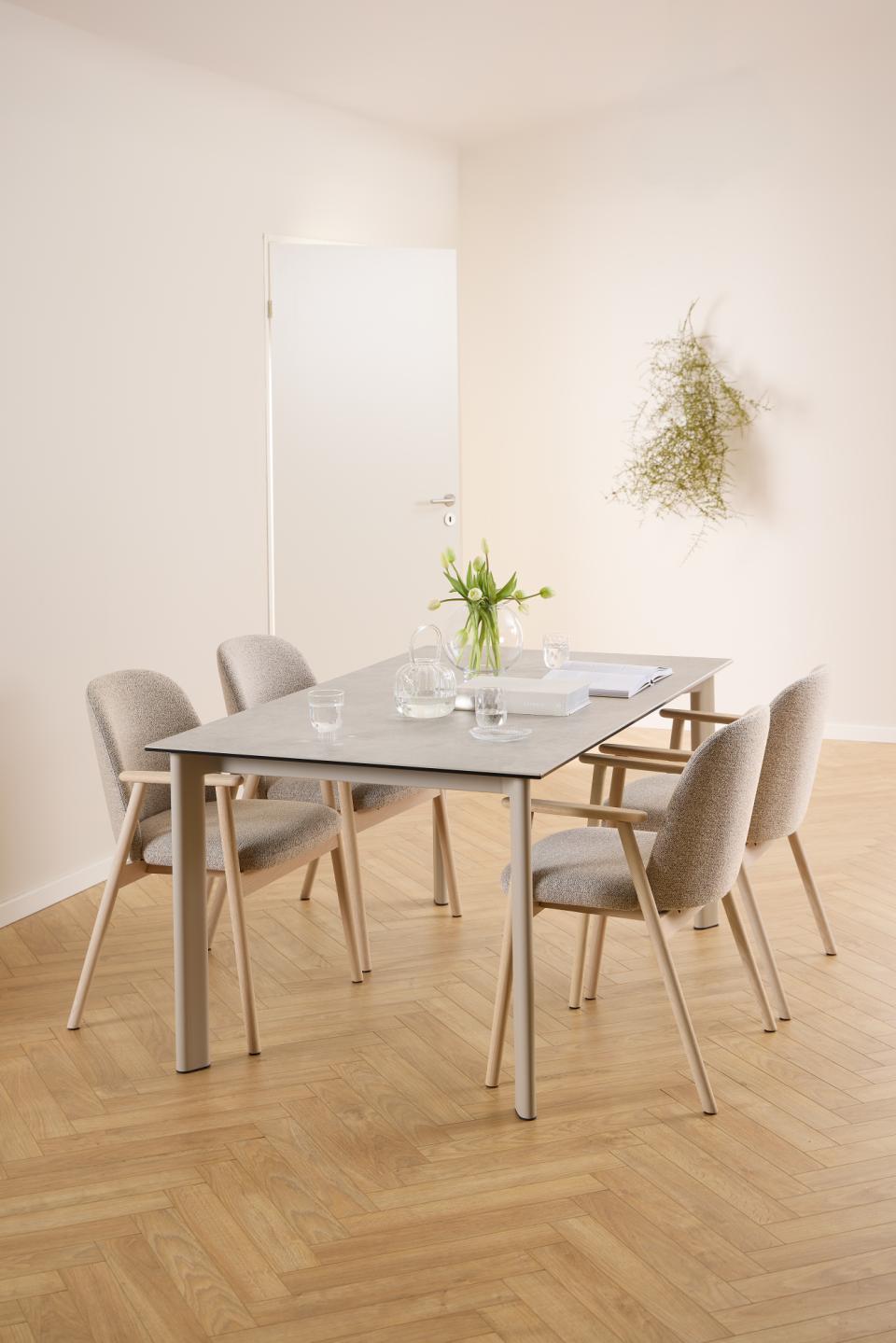A modern dining set featuring a rectangular gray stone-top table with light wood legs, surrounded by six upholstered dining chairs in a neutral beige tone, set in a minimalist dining room with light wood herringbone flooring.