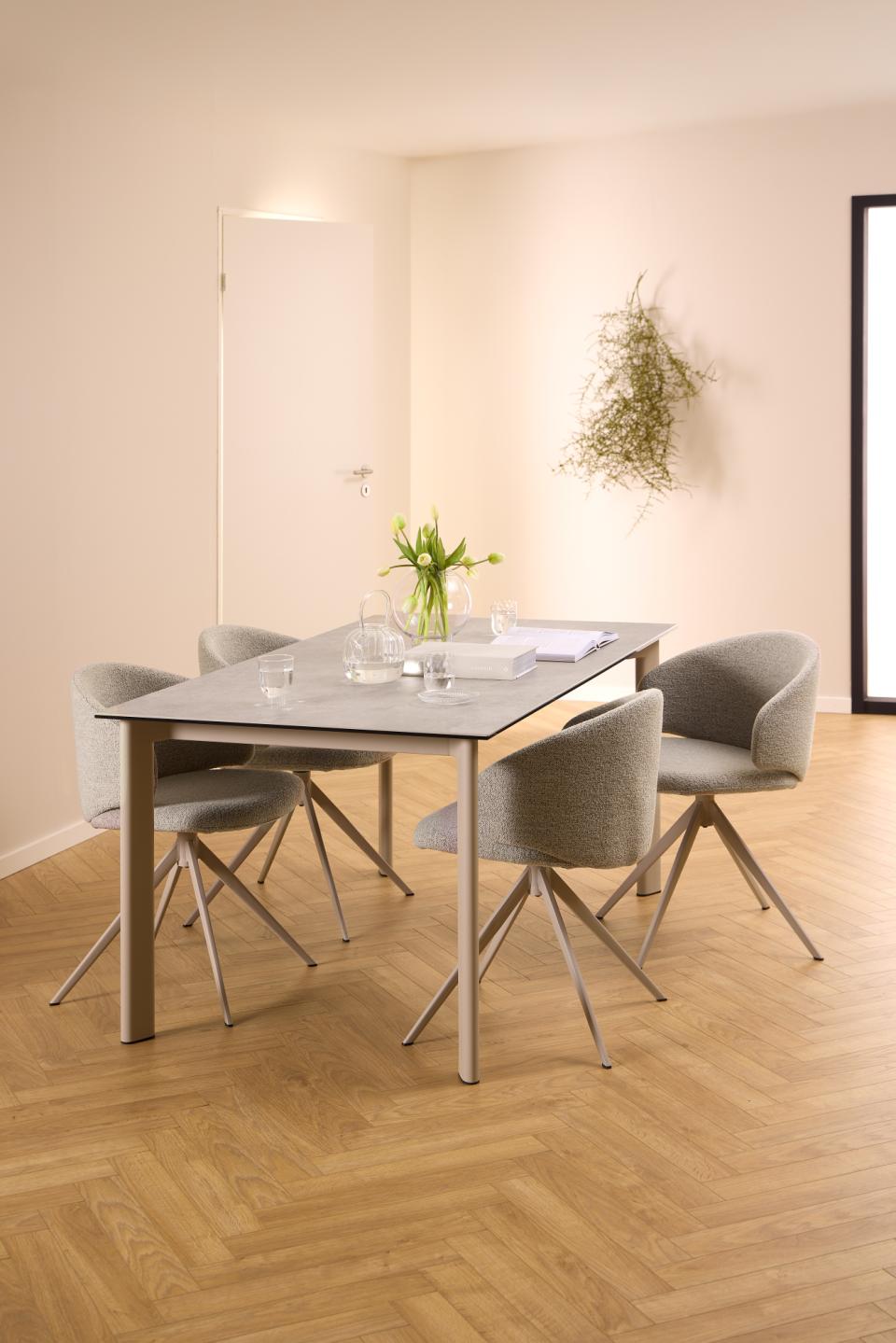 A modern dining set featuring a rectangular gray stone-look table with a light wood base and four gray upholstered chairs with metal legs, set in a minimalist dining room with beige walls and herringbone wood flooring.