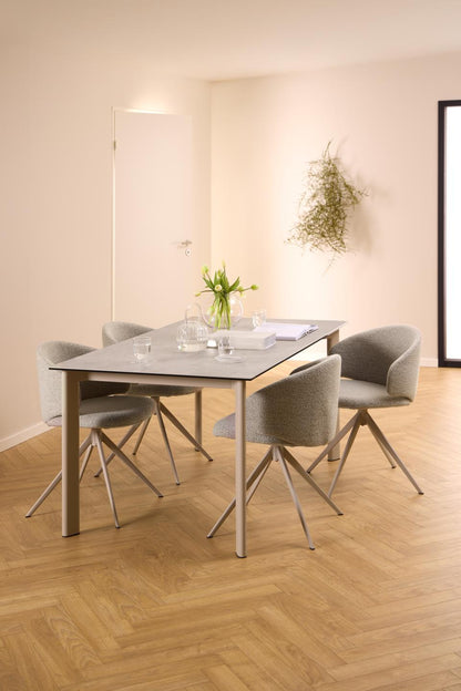 A modern dining set featuring a rectangular gray stone-look table with a light wood base and four gray upholstered chairs with metal legs, set in a minimalist dining room with beige walls and herringbone wood flooring.