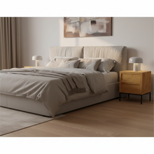 Load image into Gallery viewer, A modern upholstered bed in a warm beige fabric, styled with neutral-toned bedding and pillows, flanked by two light wood nightstands with white lamps, set against a minimalist beige wall with a piece of abstract wall art.
